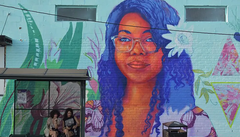 Two individuals are sitting at a bus stop in front of a colorful mural of a woman with blue hair painted on a buildings wall