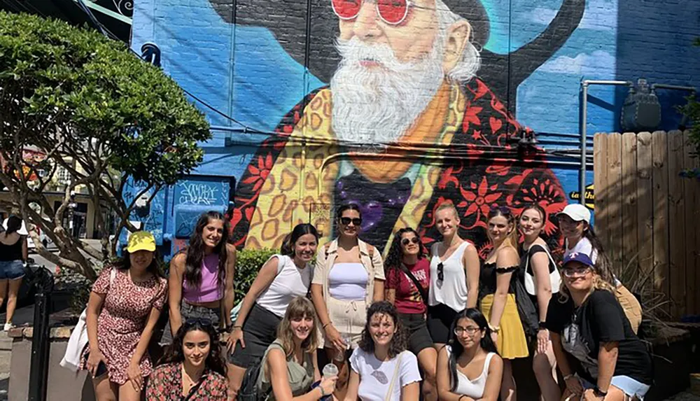 A group of people is posing for a photo in front of a colorful mural featuring a large depiction of an elderly man with a beard and sunglasses