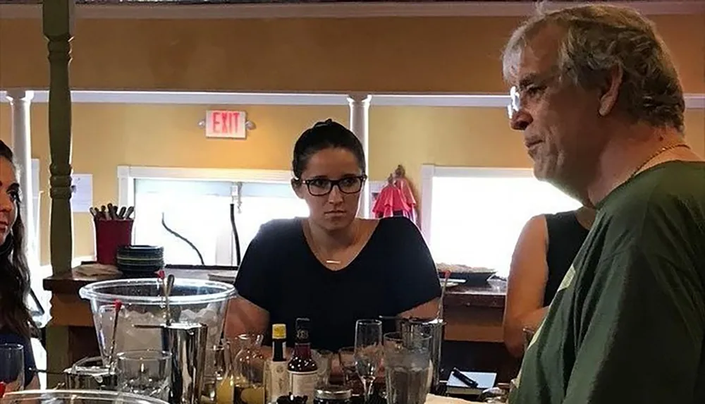 A woman with glasses appears to be listening intently or thinking with a somewhat stern expression while a man is seen in profile in the foreground all set in a casual dining environment with various glassware on the table