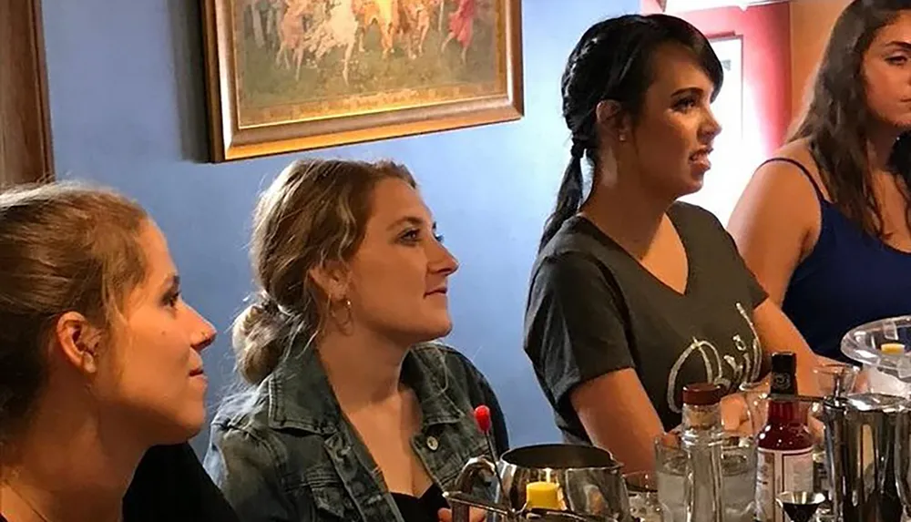 Four women are attentively watching something off-camera at a table with various drinkware and condiments suggesting they are in a dining setting