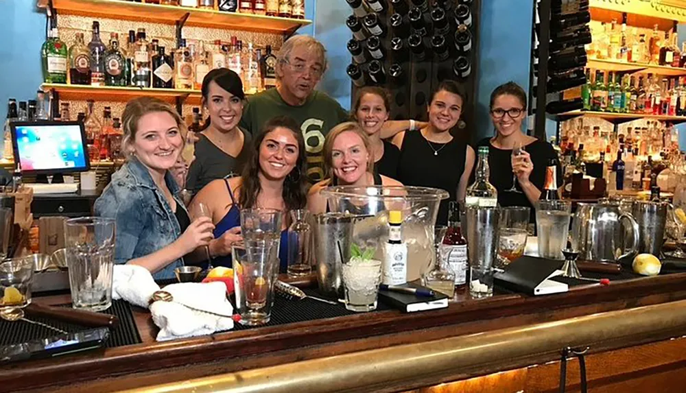 A group of people is posing with smiles behind a bar counter cluttered with cocktail-making equipment and glasses