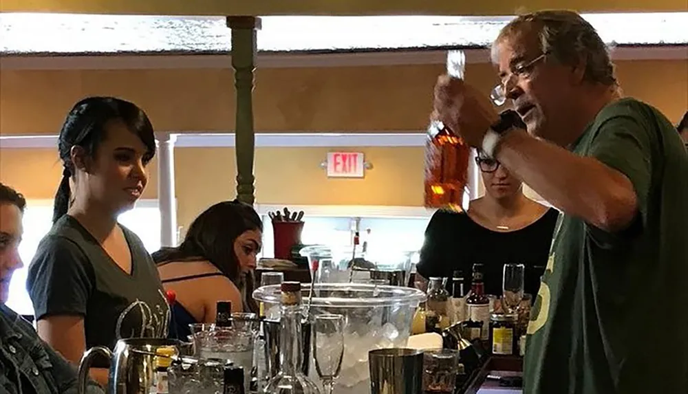 A man is pouring a drink at a bar where several people are gathered with one woman in the foreground seemingly glancing at the camera