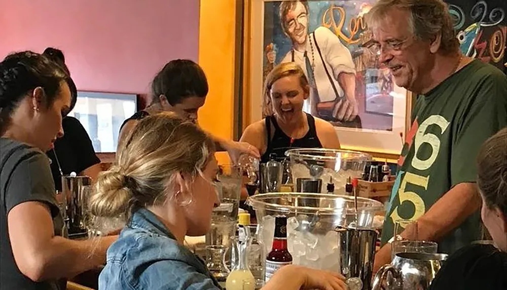 A group of people appears to be enjoying a casual cocktail mixing class or social gathering with drinks featuring a cheerful gentleman in a green shirt engaging with others around a table filled with glassware and beverage-making ingredients