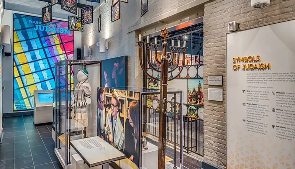 This image depicts an exhibit highlighting Symbols of Judaism featuring a menorah a tallit on a mannequin educational panels and a colorful stained glass window