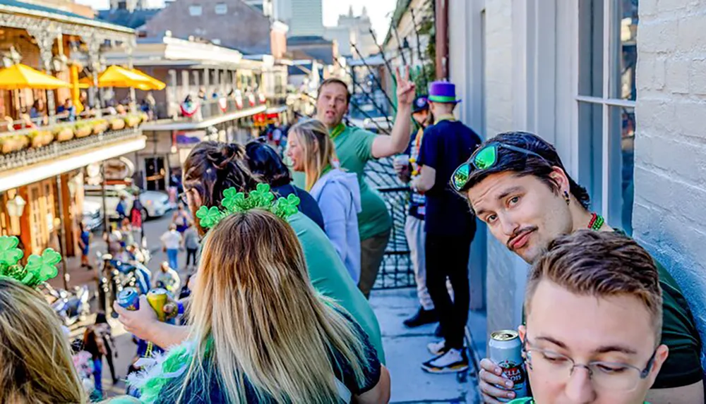 A group of people wearing green accessories and holding drinks appear to be enjoying a festive outdoor event likely a St Patricks Day celebration on a balcony overlooking a busy street