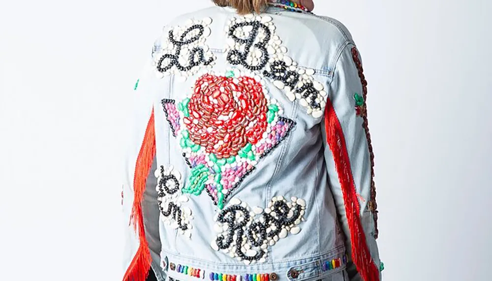 A person is wearing a heavily embellished denim jacket with red fringe beads and a large rose design on the back