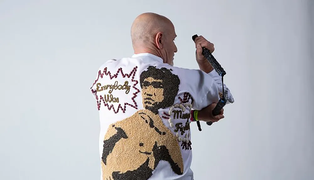 A bald person is viewed from behind wearing a white shirt adorned with a beaded image of a man and a horse alongside the phrase Everybody Was Kung Fu Fighting