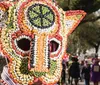 The image shows a person wearing a large elaborate mask or costume headpiece made of colorful beans set against the backdrop of an outdoor celebration with people in the background suggesting a festive or cultural event