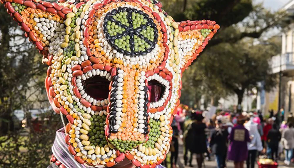 The image shows a person wearing a large elaborate mask or costume headpiece made of colorful beans set against the backdrop of an outdoor celebration with people in the background suggesting a festive or cultural event