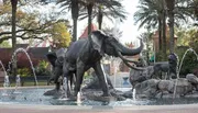 The image captures a dynamic fountain display featuring statues of elephants and lions surrounded by water jets, set in a serene landscape with palm trees and a clear sky.