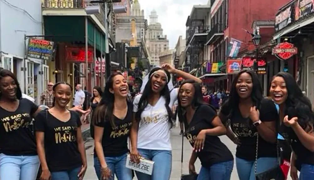 A group of women is joyously walking down a vibrant street wearing matching t-shirts that suggest a celebratory event possibly a bachelorette party given the sash and tiara worn by the woman in the center