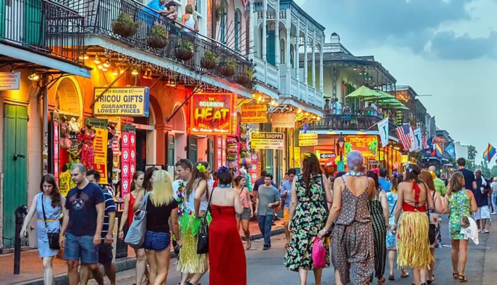 A vibrant crowd strolls along a colorful and busy Bourbon Street in New Orleans flanked by ornate balconies and neon signs as dusk sets in