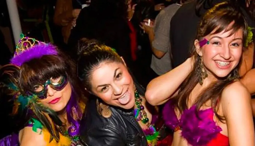 Three people at a festive gathering are smiling and posing for the camera with one of them wearing a masquerade mask and a tiara suggesting a costume or celebratory event