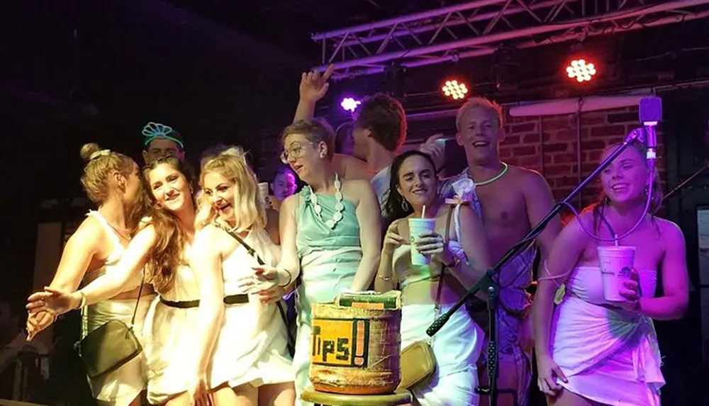 A group of people dressed in white and toga-style outfits are having fun on a stage with drinks in their hands and a Tips jar in front of them indicating a lively party or social event