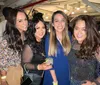 Four women are smiling for a photo at a social gathering with one holding a beverage