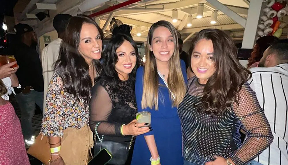 Four women are smiling for a photo at a social gathering with one holding a beverage