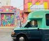 A colorful mural with vibrant mythical creatures adorns a building behind a teal-colored bus on a wet street