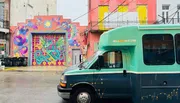 A colorful mural with vibrant mythical creatures adorns a building behind a teal-colored bus on a wet street.