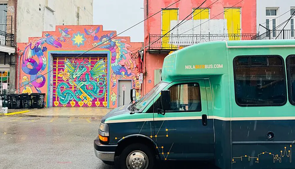 A colorful mural with vibrant mythical creatures adorns a building behind a teal-colored bus on a wet street