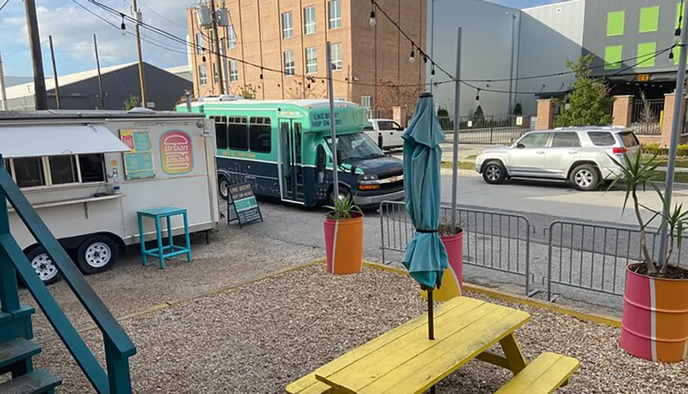 The image depicts a casual outdoor setting with a food truck parked beside a seating area featuring a yellow picnic table colorful planters and folded umbrellas inviting a street-food dining experience