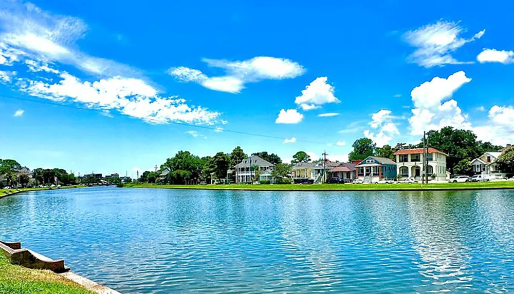 The image shows a serene blue waterway lined with trees and houses under a bright blue sky dotted with fluffy white clouds