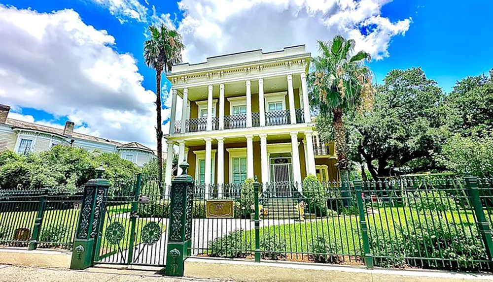 This image displays a grand two-story yellow house with white columns and balconies surrounded by a green wrought-iron fence under a blue sky with scattered clouds