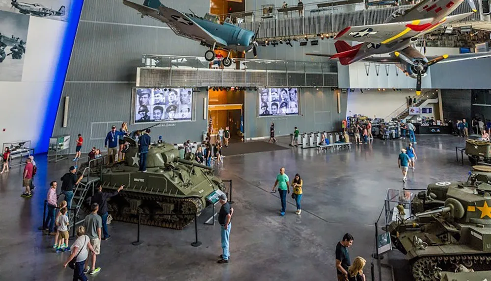 A museum exhibit features tanks on display and aircraft suspended from the ceiling with visitors exploring the area