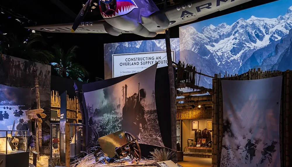 The image depicts a museum exhibit featuring a suspended aircraft with a shark face design surrounded by historical displays and scenic backdrops of mountains