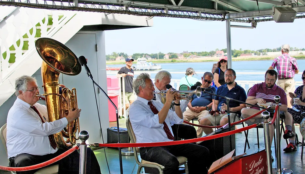 Musicians play brass instruments for an attentive audience on a boat with a scenic view in the background