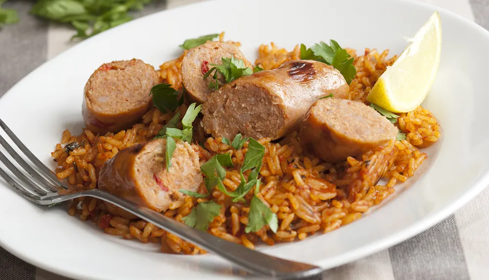 A plate of flavorful rice topped with sliced sausage garnished with fresh herbs and a lemon wedge