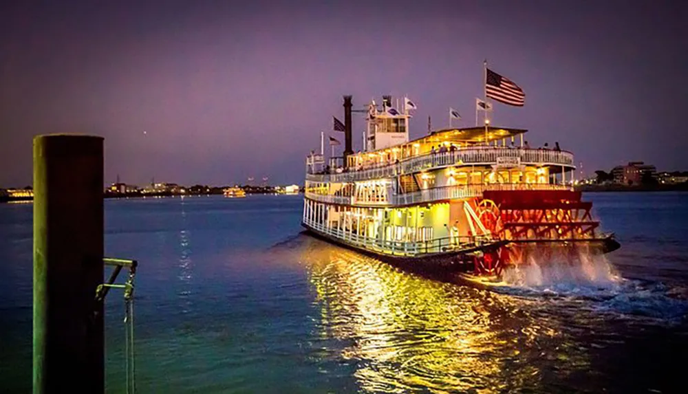 A brightly lit paddle steamboat cruises down a calm river under a dusky sky