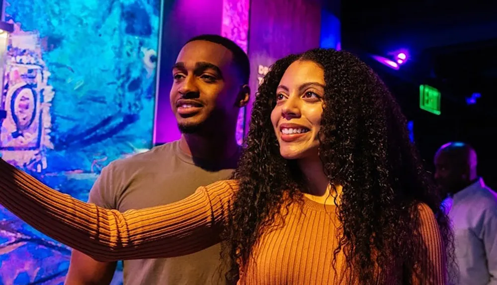 A couple is smiling and looking at something in a colorful dimly lit environment