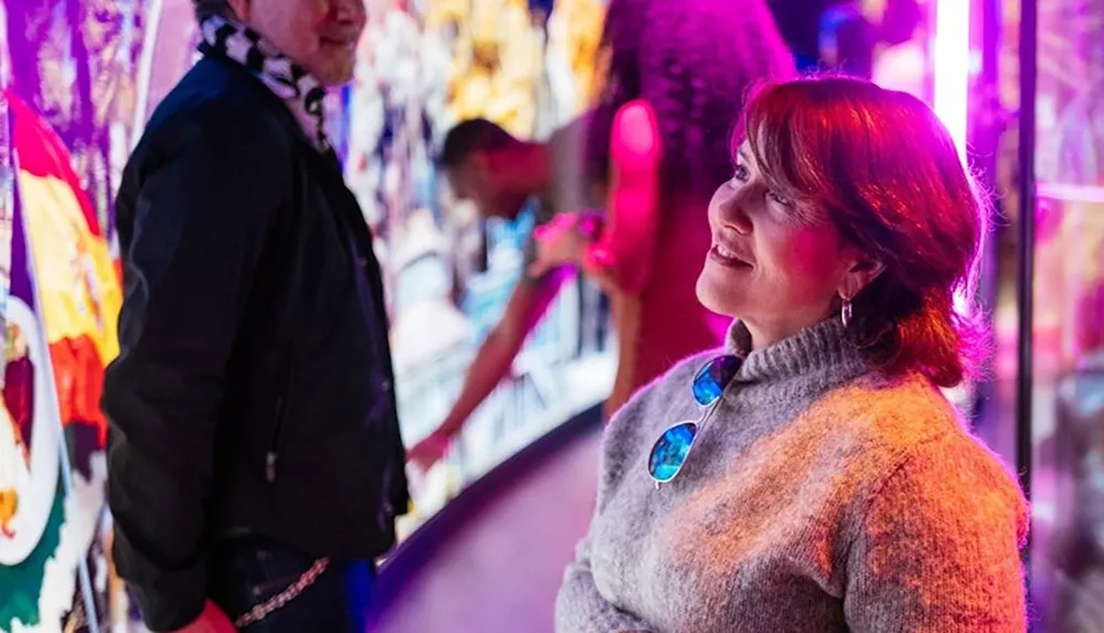 A woman in a cozy sweater is smiling illuminated by vibrant purple and pink lights with a colorful backdrop and people around her