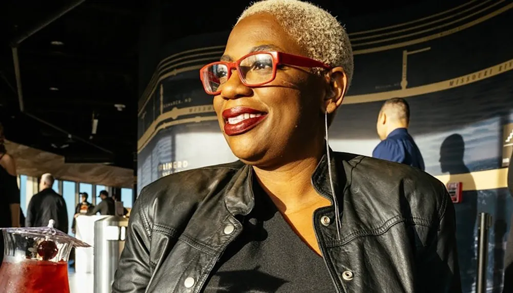 A person with short blonde hair and red glasses is smiling while wearing a black leather jacket in a stylish indoor setting