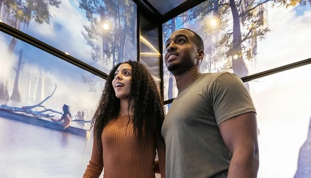 A couple stands in front of a scenic display featuring a serene forested lake looking awestruck and engaged