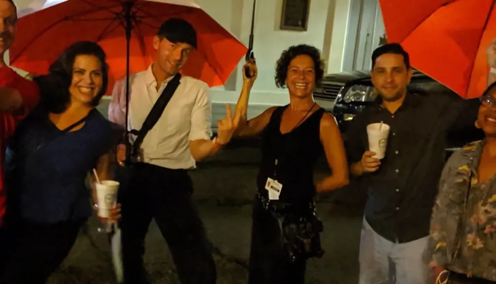 A group of people are happily posing with red umbrellas some holding drinks on a street at night