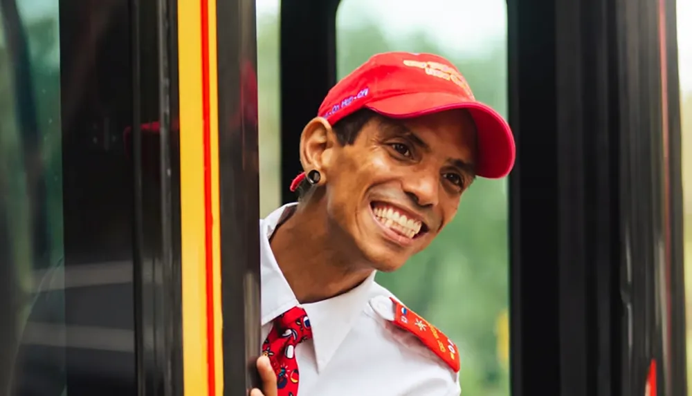 A smiling person wearing a red cap and uniform leans out of a bus door