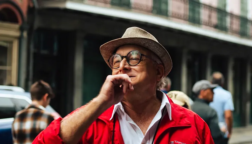 A smiling man in a red jacket and straw hat stands on a street surrounded by people in the background