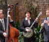 A trio of musicians play instruments in an outdoor setting with an inset of a table featuring a plate of food and a drink