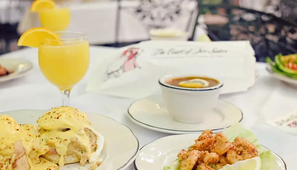 The image depicts a table setting with a glass of orange juice a plate of eggs Benedict a bowl of soup and a dish of shrimp accompanied by a Court of Two Sisters menu in the background