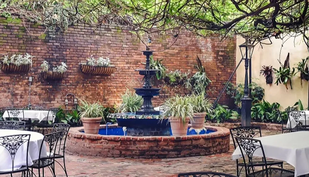 The image depicts a charming courtyard with a decorative fountain surrounded by plants and tables with white tablecloths under a canopy of greenery