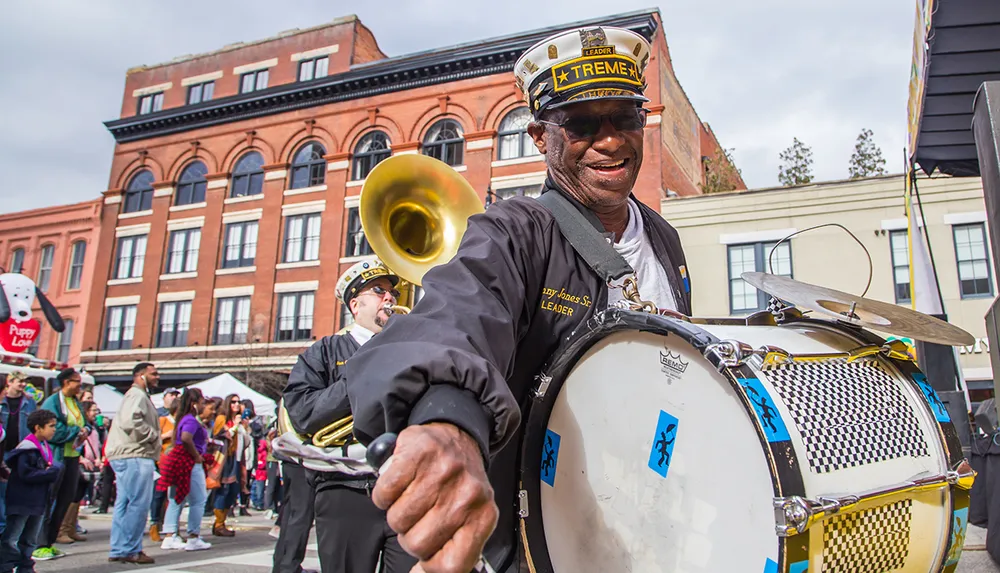 A smiling man in a marching band uniform plays a drum in a lively street parade