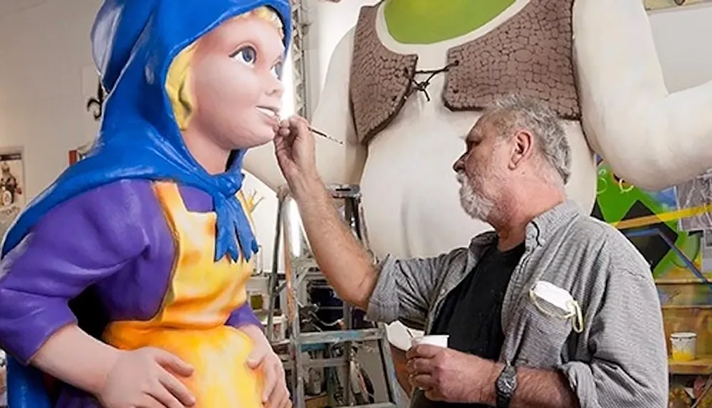 An artist is painting a colorful sculpture of a character wearing a blue hood with another large figure visible in the background