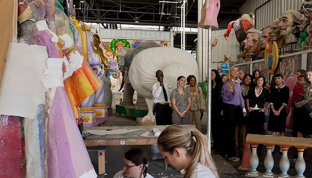 A group of people is observing artists working in a workshop filled with large colorful sculptures and props