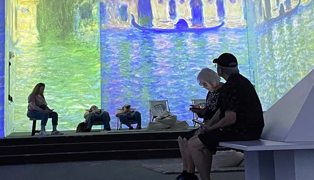 People are sitting on benches and deck chairs in a room illuminated by large projections of impressionist art