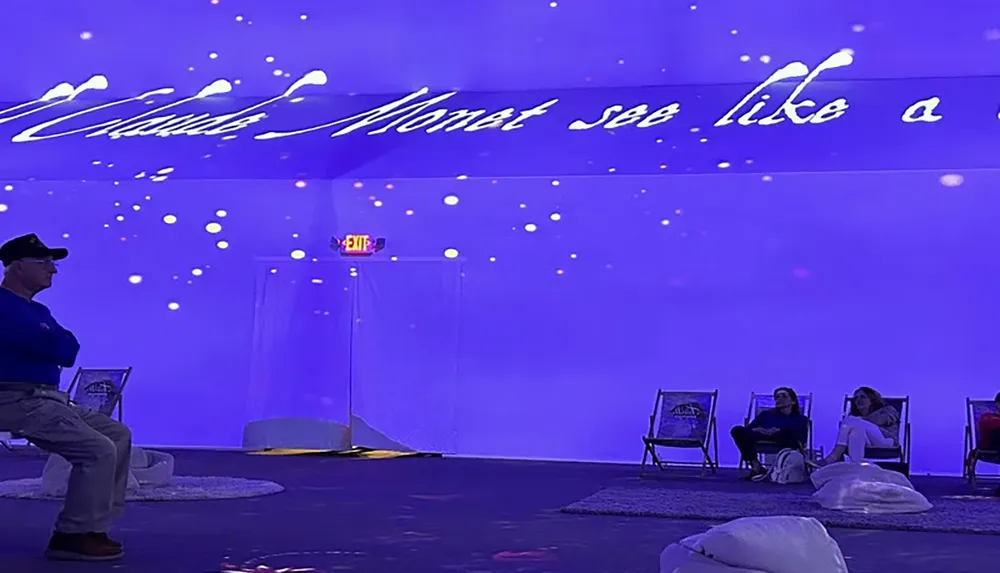 The image shows a room with a blue-lit immersive art exhibit where people are seated on chairs with the words Claude Monet see like a projected on the wall