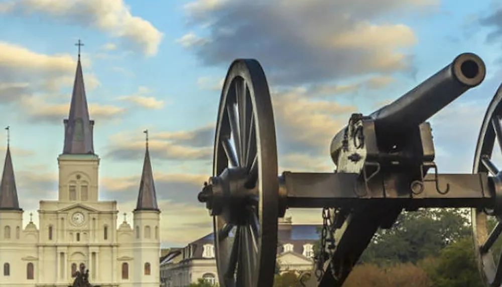 A historic cannon is positioned in the foreground with a grand church featuring tall spires set against a partly cloudy sky