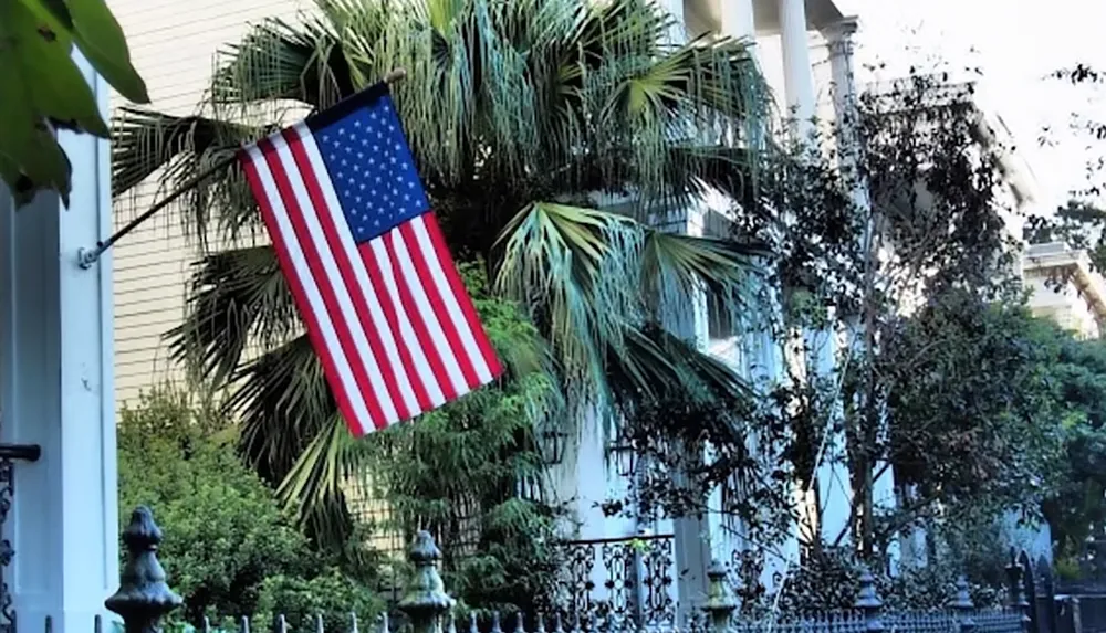 A United States flag is hanging in front of a house surrounded by lush palm trees and greenery