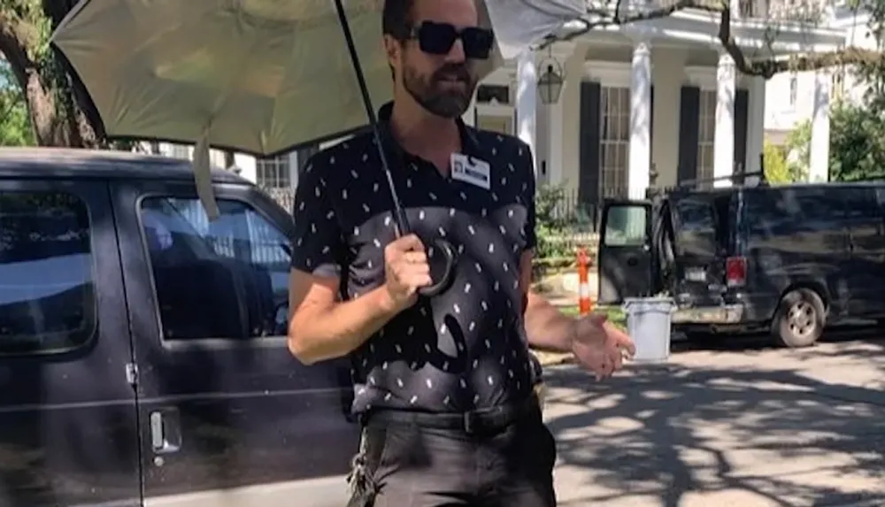 A person wearing a patterned shirt and sunglasses holds an umbrella while standing near a dark van on a sunny day