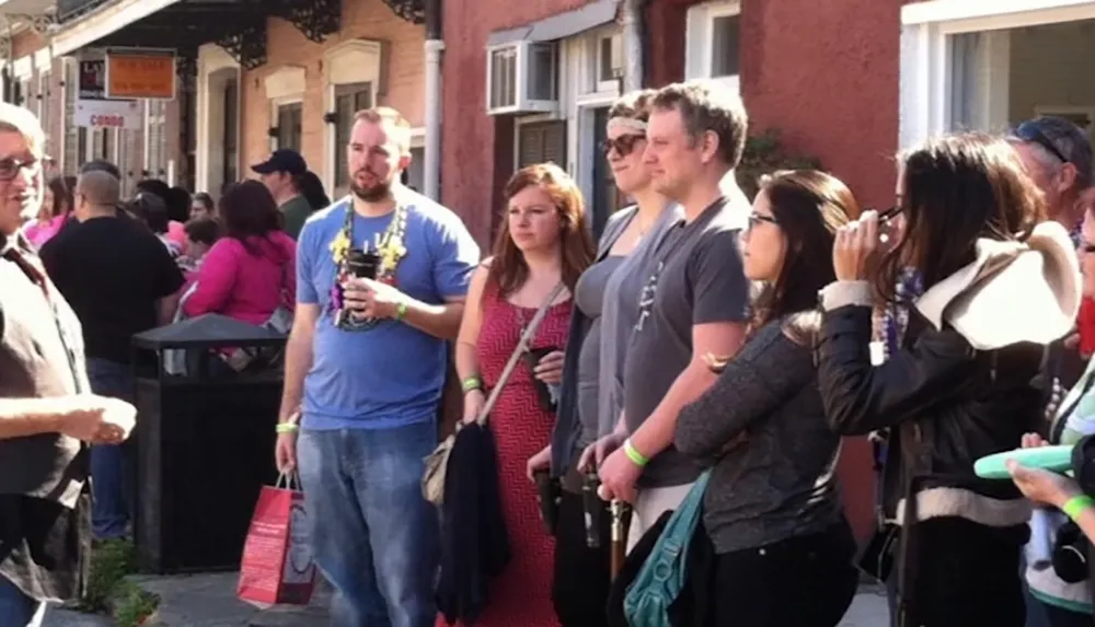 A group of people is casually standing in a line on a busy street some holding drinks and wearing casual attire